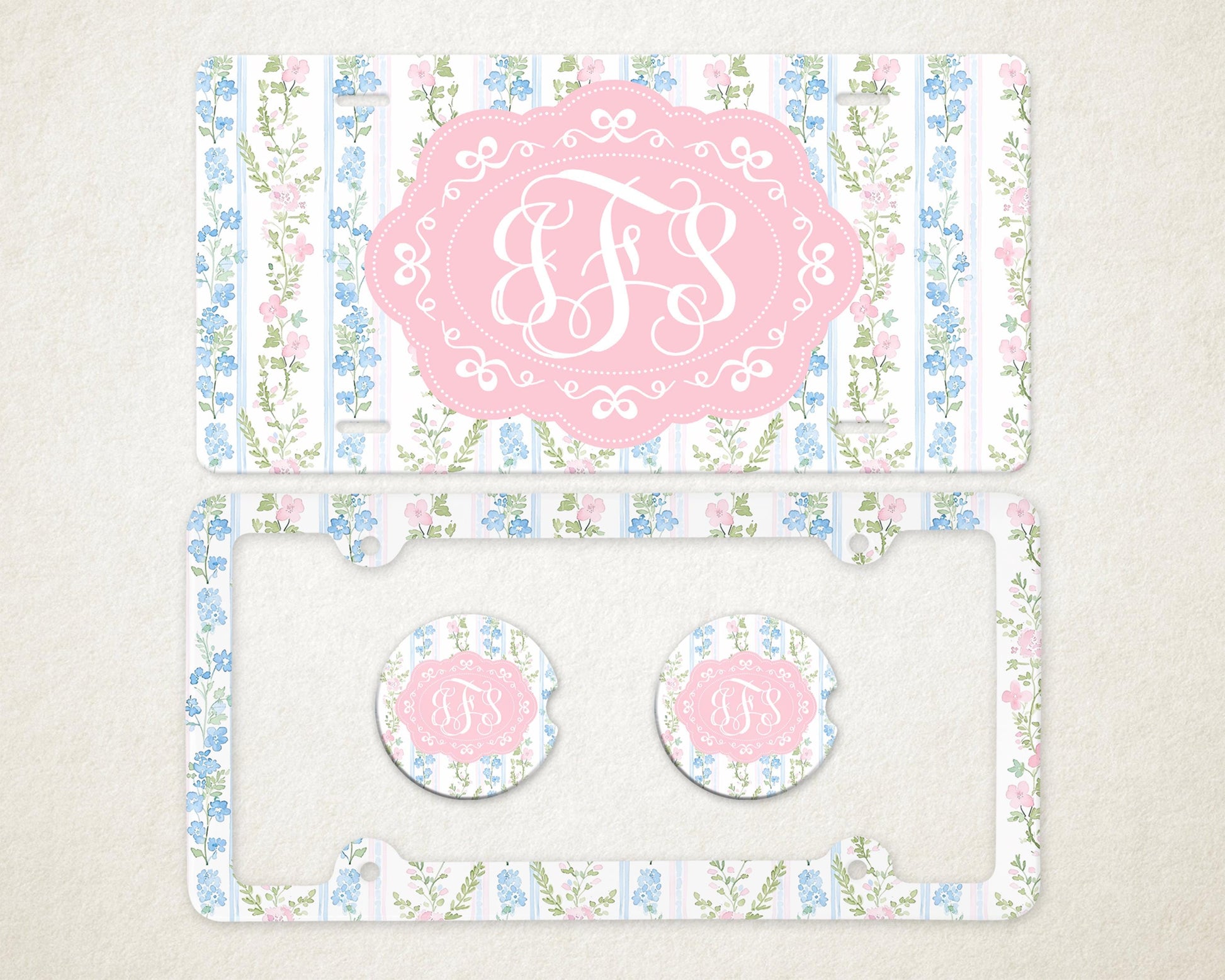 May include: A license plate, license plate frame, and two car coasters with a floral design. The items feature a pink and white monogram in a pink oval frame, set against a background of blue and pink flowers.