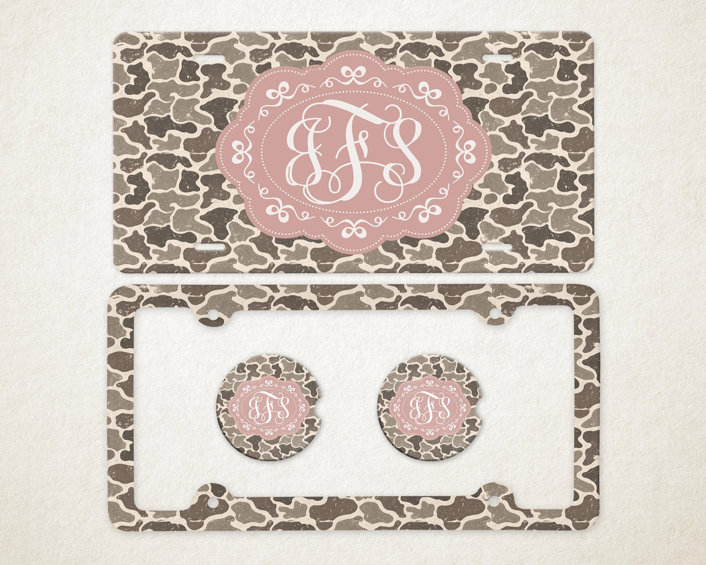 May include: A license plate, license plate frame, and two car coasters with a camouflage pattern in shades of brown and gray. Each item features a pink oval with a white monogram.
