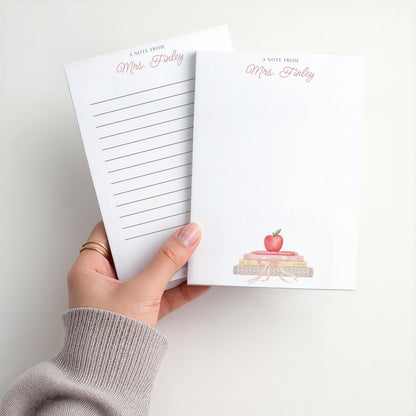 May include: White notepad with the text "A Note From Mrs. Finley" in pink script. The notepad features a watercolor illustration of a stack of books topped with a red apple. One notepad has lined pages.